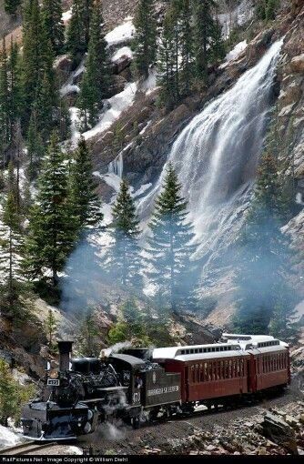 a train traveling down tracks next to a forest covered hillside with a waterfall in the background