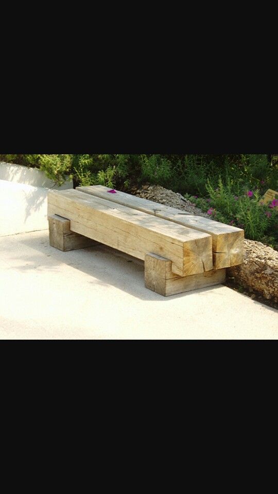 a wooden bench sitting on top of a cement ground next to trees and bushes with purple flowers in the background