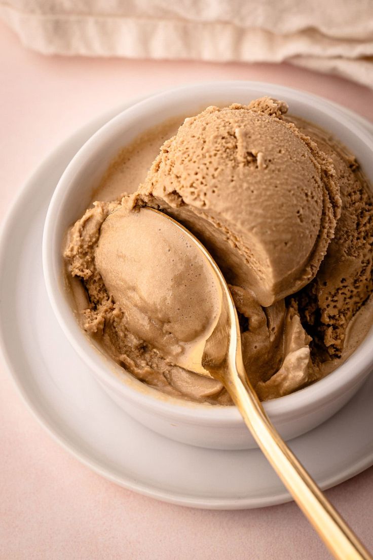 a white bowl filled with ice cream on top of a pink surface next to a gold spoon