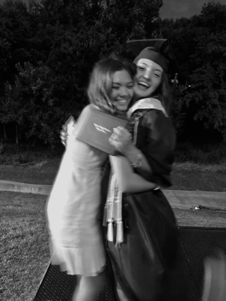 two women hugging each other in front of some trees and grass with one holding a book