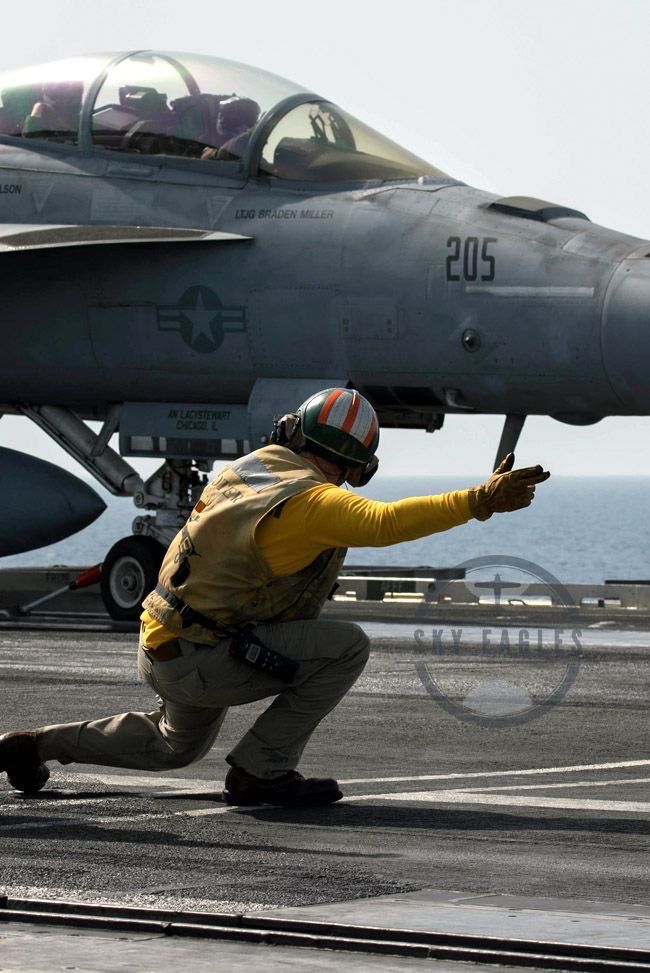 a fighter jet sitting on top of an aircraft carrier next to a man in yellow