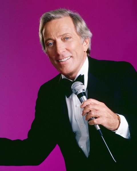 a man in a tuxedo holds a microphone and points to the side with his right hand