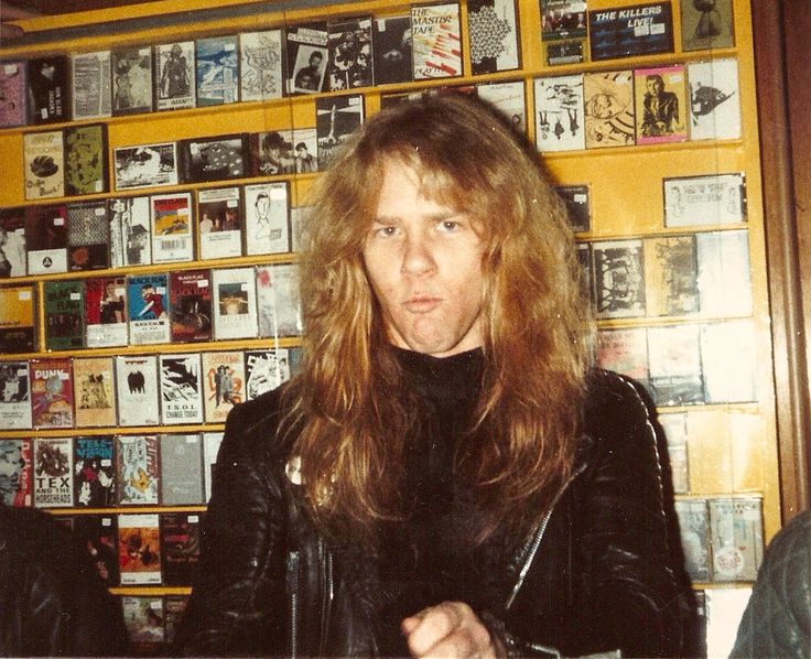 a man with long hair standing in front of a wall full of books and magazines