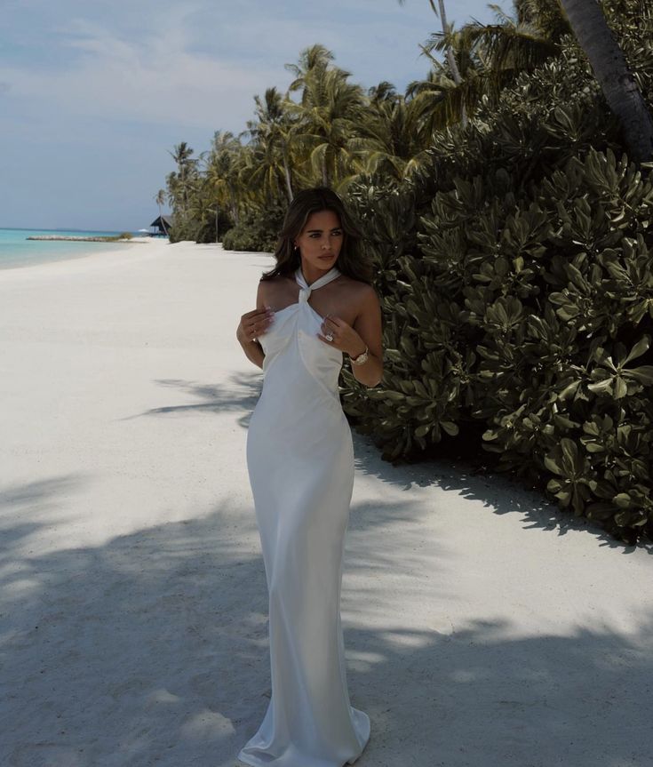 a woman in a white dress standing on the beach next to some trees and bushes