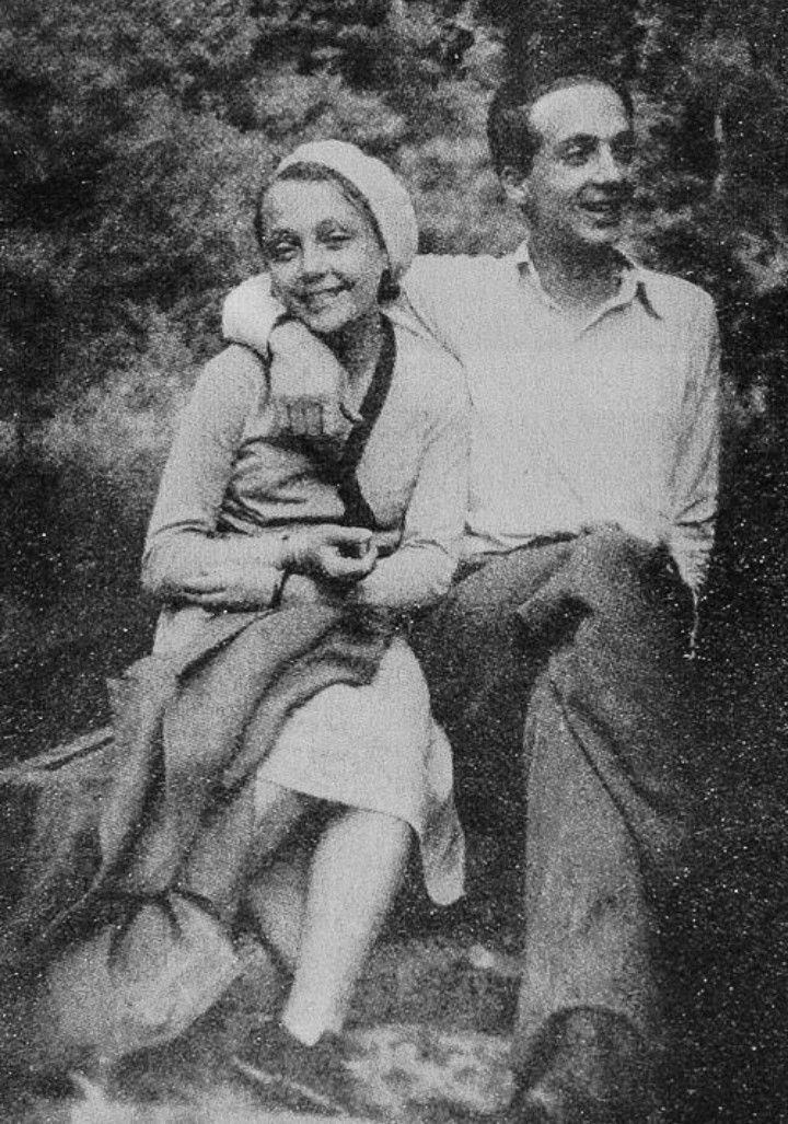 an old black and white photo of a man and woman sitting on a rock in the woods