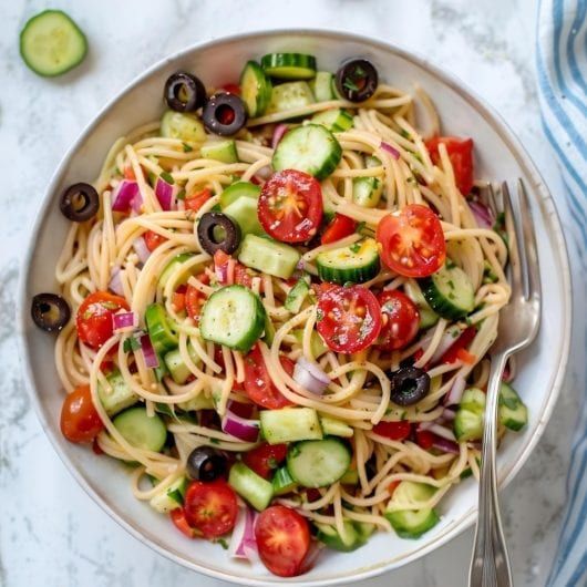 a white bowl filled with pasta, cucumbers and tomatoes on top of a marble table