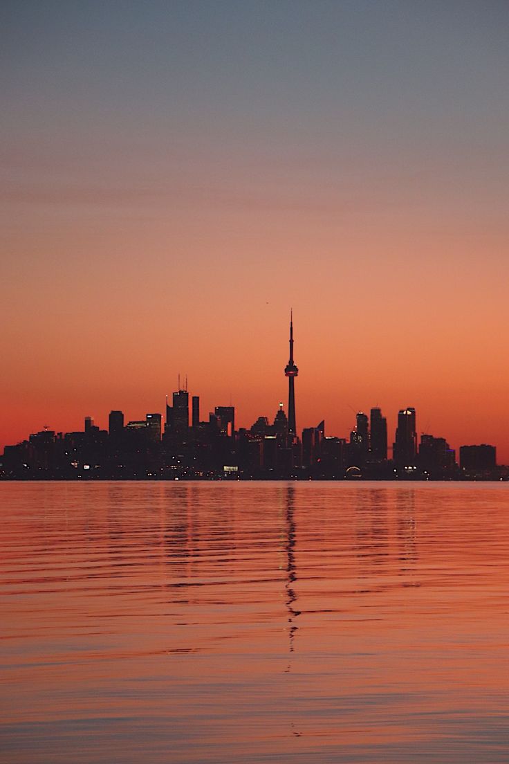 the city skyline is reflected in the calm water at sunset, with an orange glow on the horizon