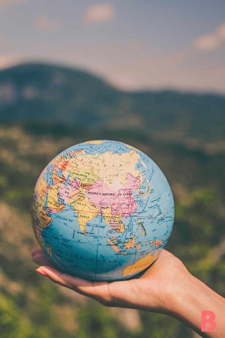 a person's hand holding up a small blue and yellow globe in front of mountains