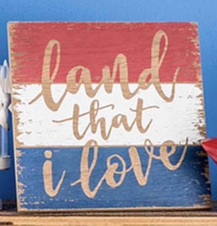 a wooden sign that says land that i love on top of a shelf next to a red white and blue flag