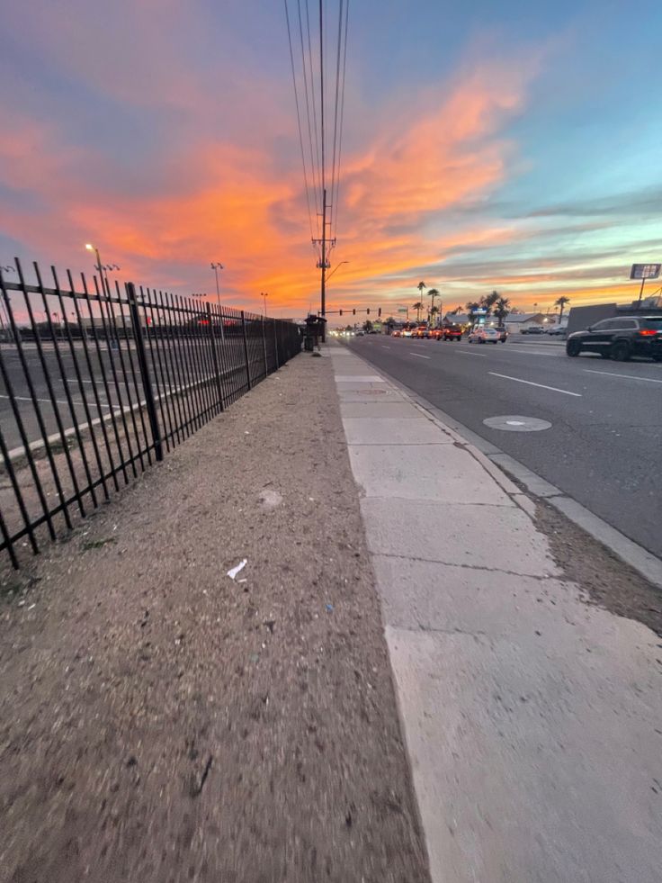 an empty sidewalk next to a fence with the sun setting in the sky behind it