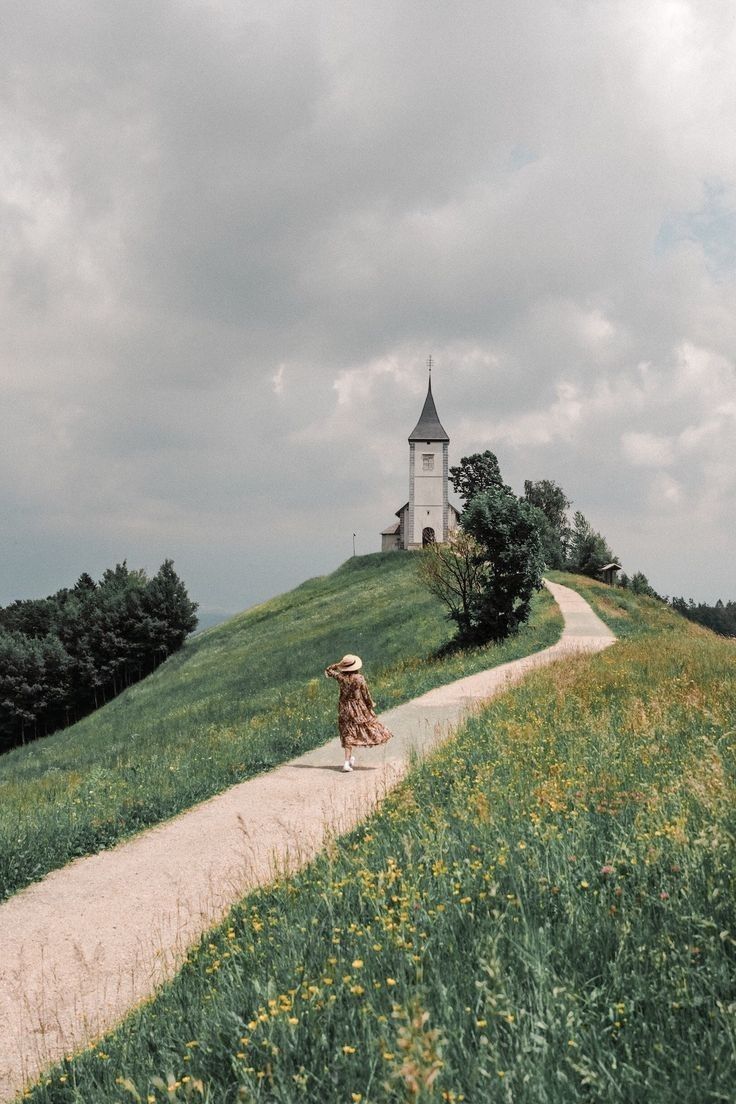 a woman walking down a dirt road towards a church on top of a grassy hill