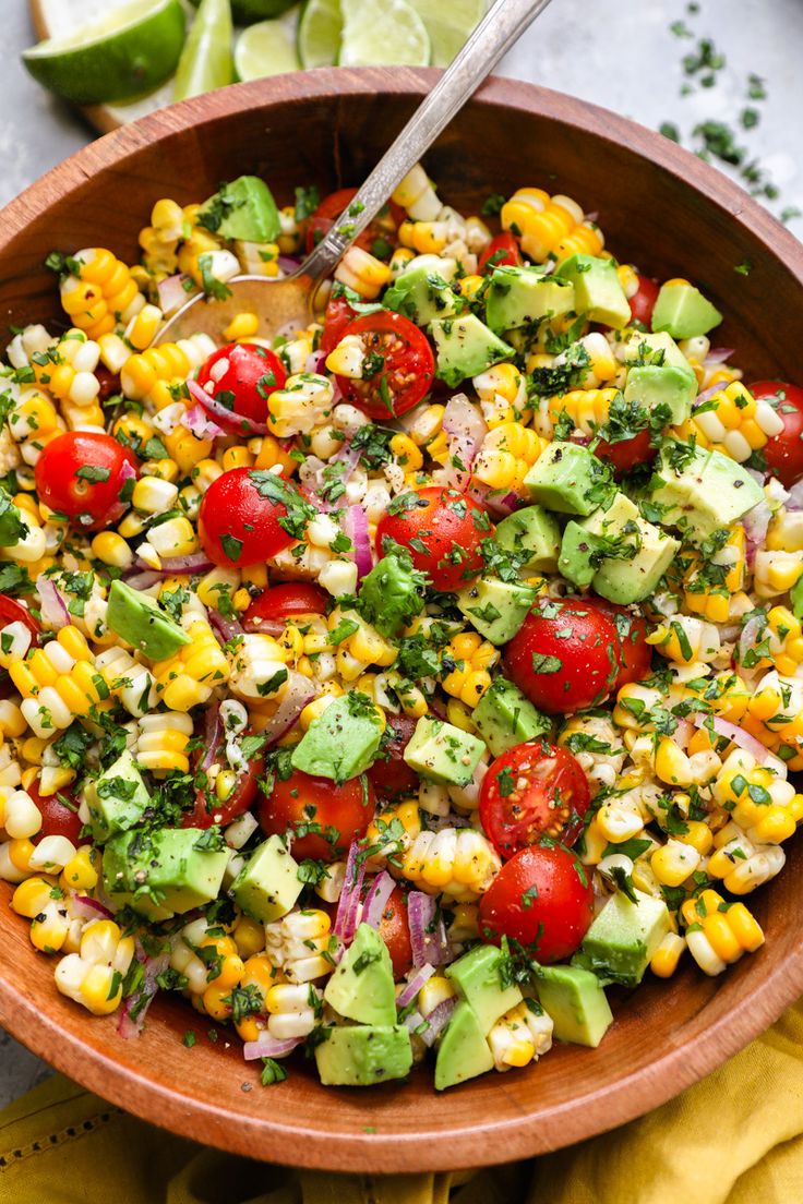 a wooden bowl filled with corn, avocado and tomato salad next to lime wedges