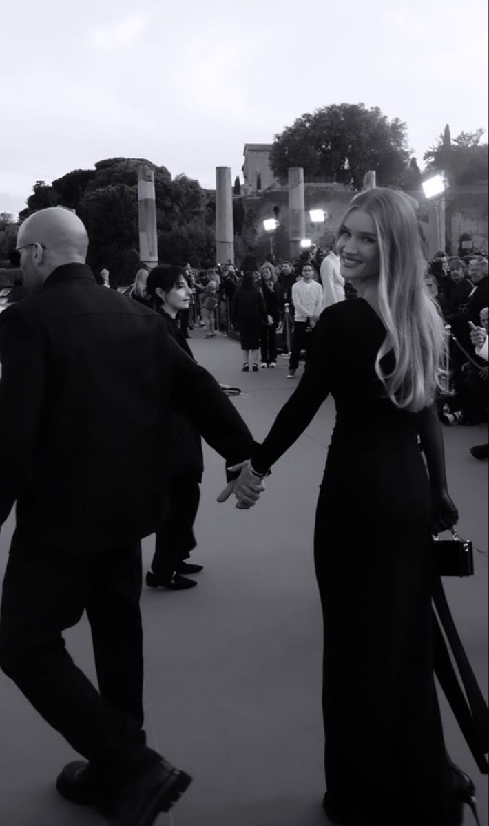 a man and woman holding hands while walking down a street with people in the background