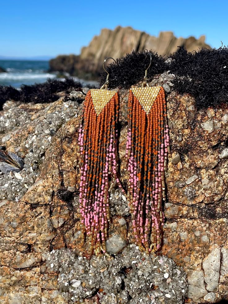 two orange and pink beaded earrings sitting on top of a rock next to the ocean