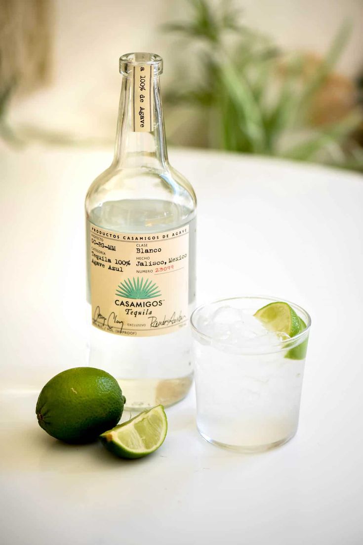 a bottle of alcohol next to a glass with limes on the table in front of it