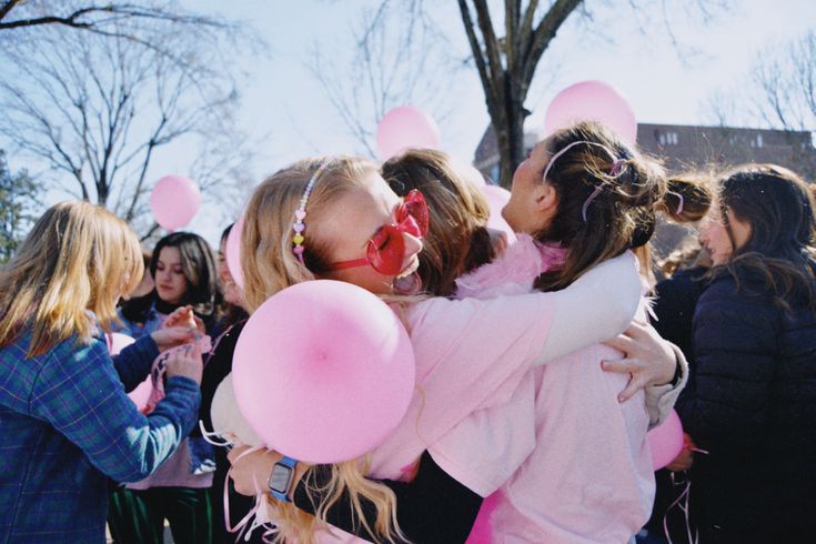 two girls hugging each other with pink balloons in the air and people standing around them