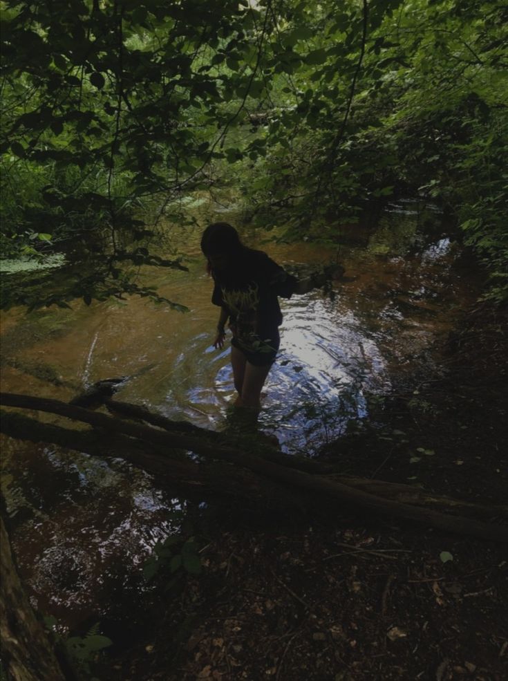 a woman standing in the middle of a stream surrounded by trees and water with her back to the camera