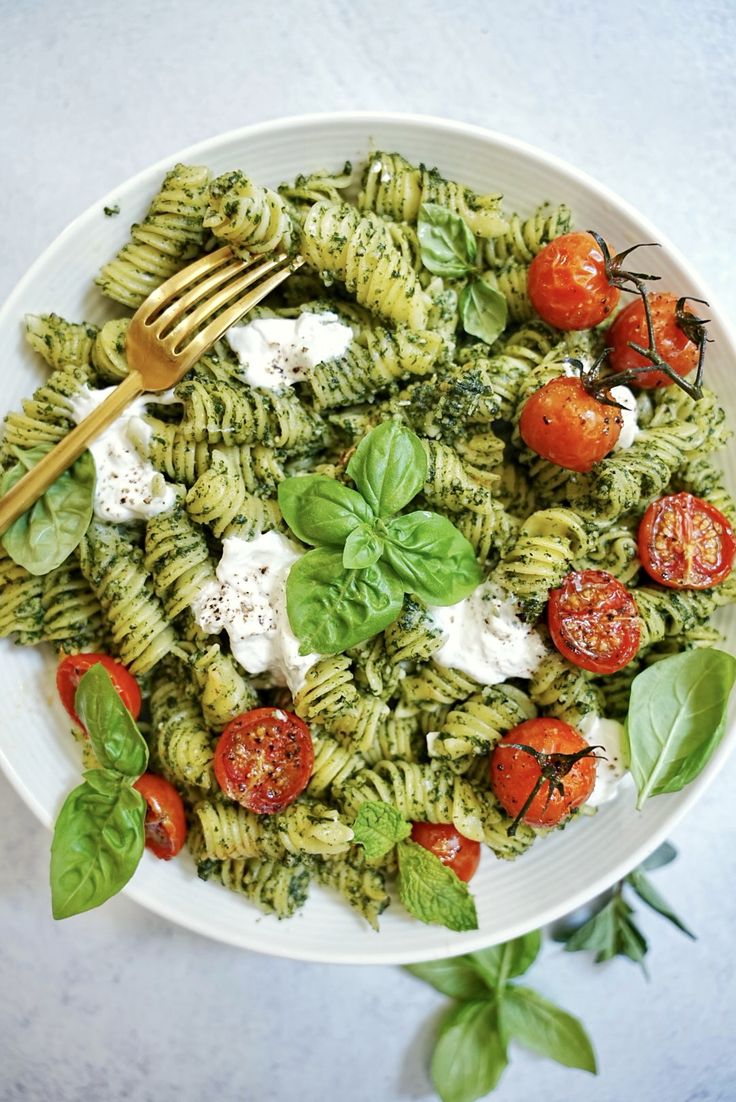 a white bowl filled with pesto pasta and topped with mozzarella, tomatoes, spinach leaves and cheese