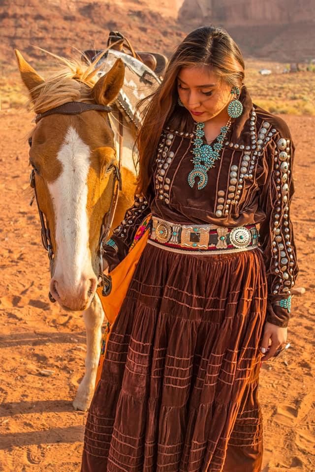 a woman in a long brown dress standing next to a horse on a dirt field