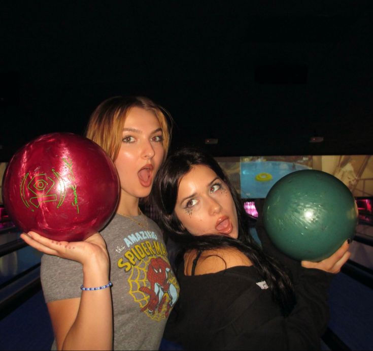 two women holding bowling balls in their hands and posing for the camera with one woman sticking out her tongue