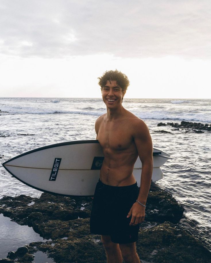 a shirtless man holding a surfboard on the rocks near the ocean at sunset