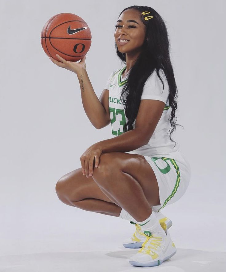 a woman holding a basketball in her right hand and sitting on the ground with one foot up