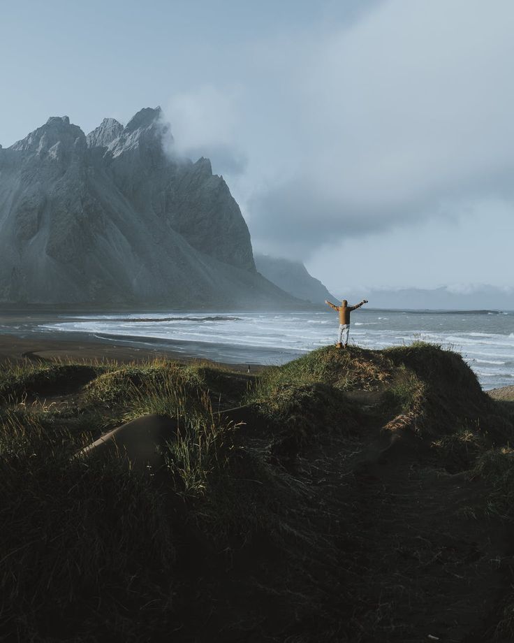 a person standing on top of a grass covered hill next to the ocean with mountains in the background