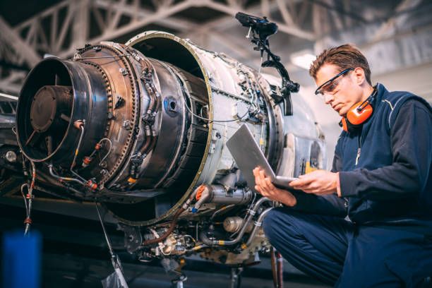 a man working on an airplane engine in a hangar with other equipment around him and looking at his tablet computer