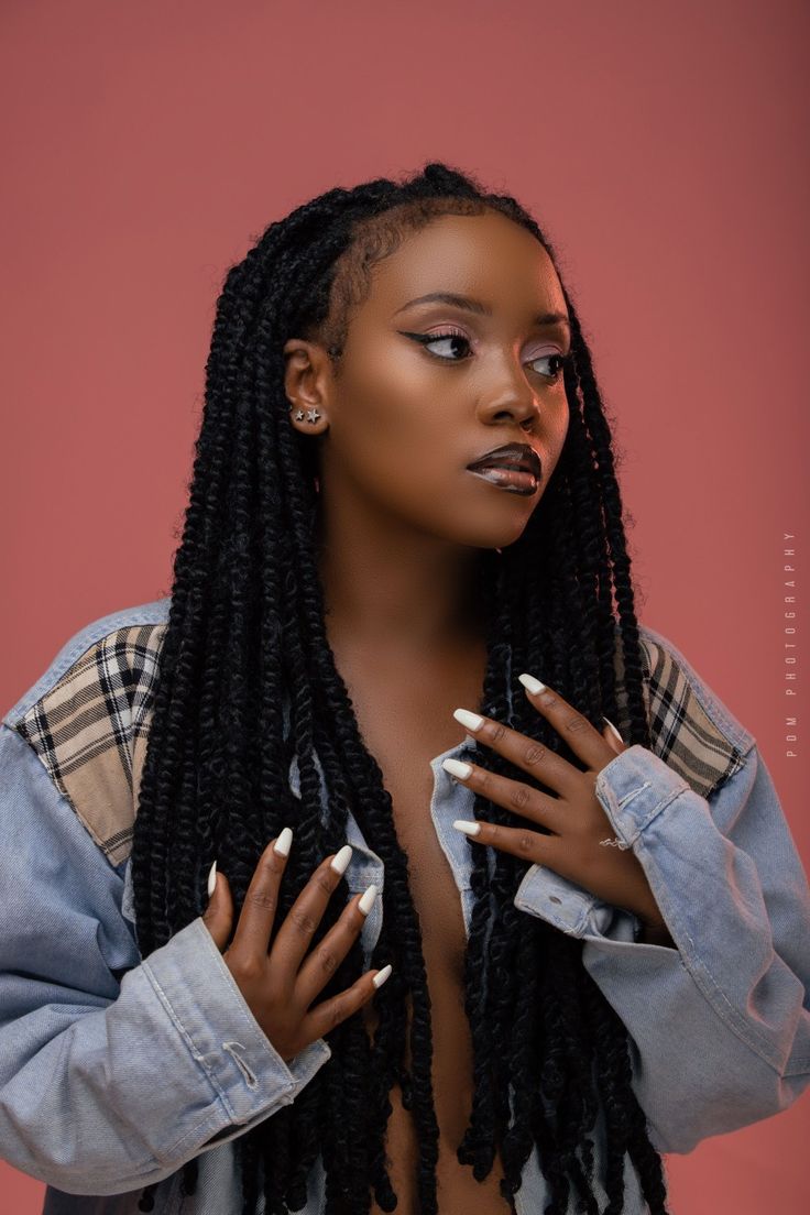 a woman with long black dreadlocks standing in front of a pink background wearing a denim jacket