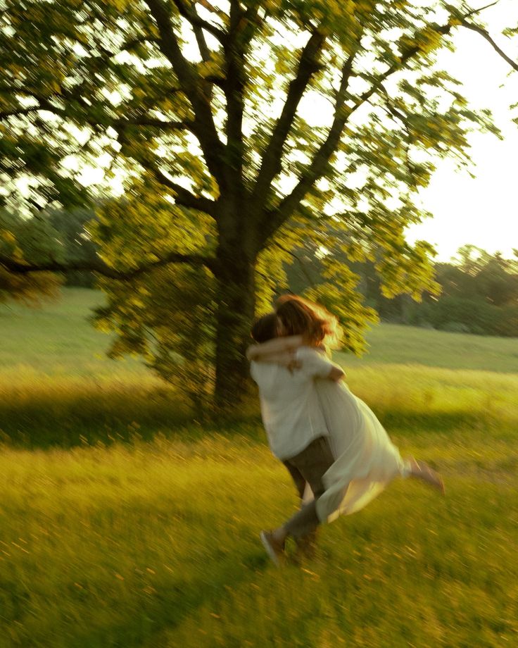 two people are playing in the grass near a tree with their arms around each other