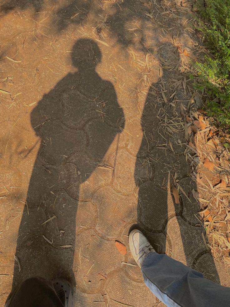 the shadow of two people standing next to each other on a dirt road with trees in the background