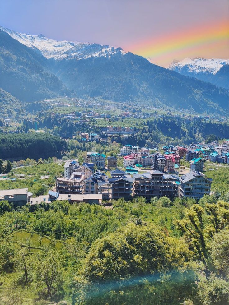 an aerial view of a city surrounded by mountains and trees with a rainbow in the sky