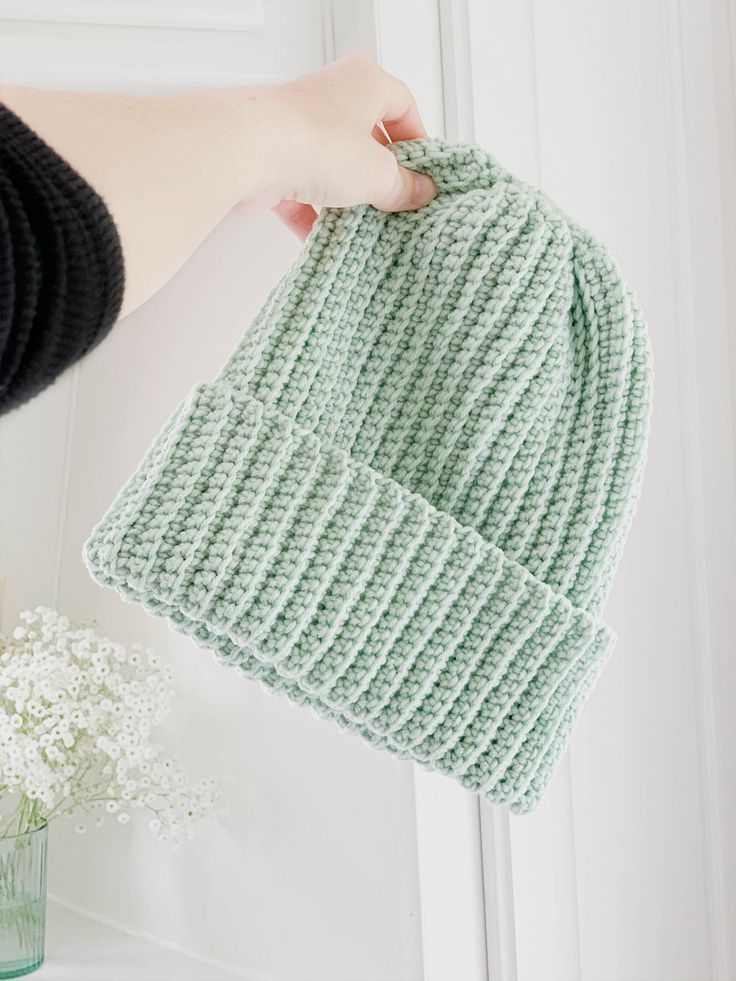 a person holding up a green knitted hat next to a vase with white flowers