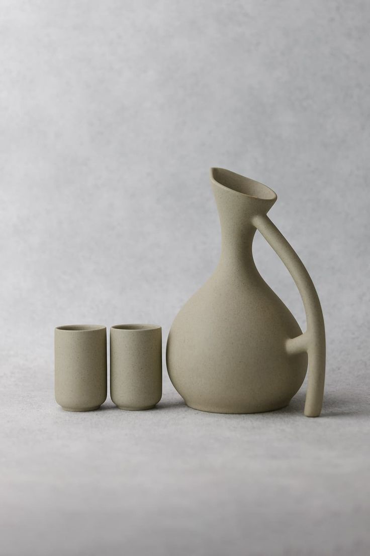 a white vase and two cups sitting next to each other on a gray surface with one empty cup in the foreground