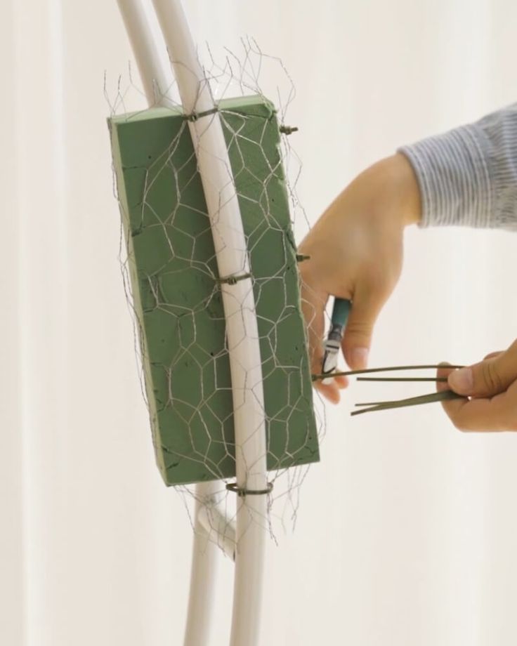 a person is cutting wire with scissors in front of a piece of green material on a white wall