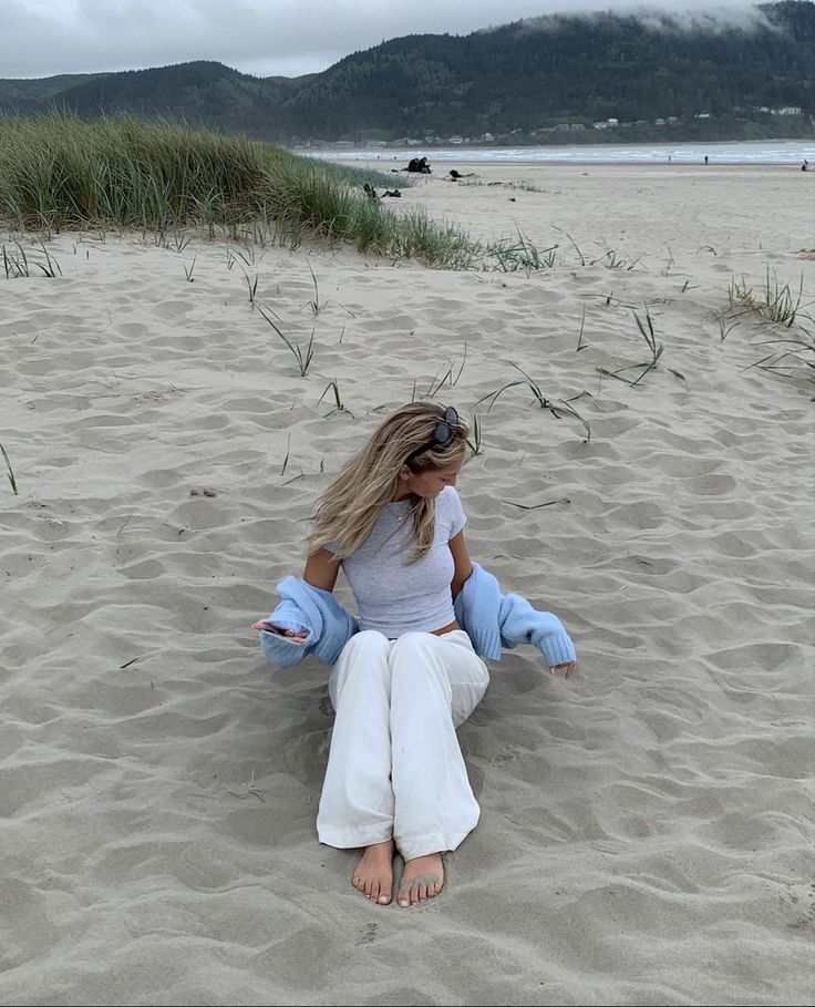 a woman sitting on top of a sandy beach next to tall grass and sand dunes