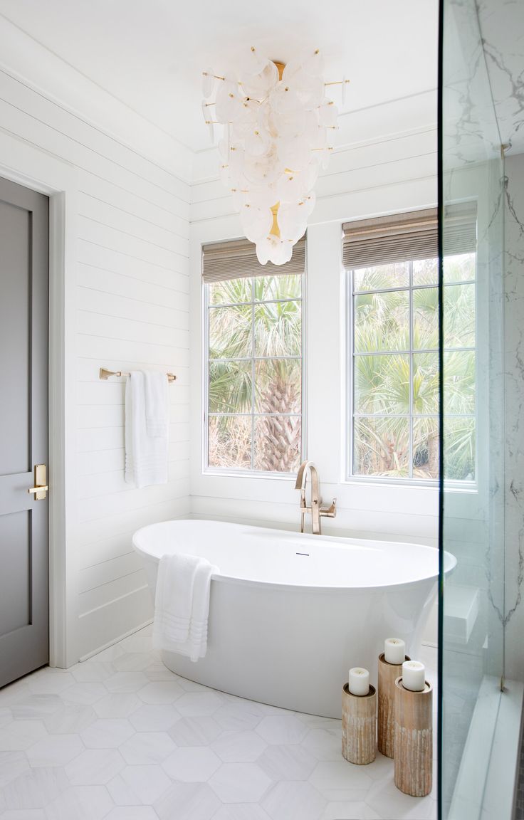 a large white bath tub sitting next to a window in a bathroom under a chandelier