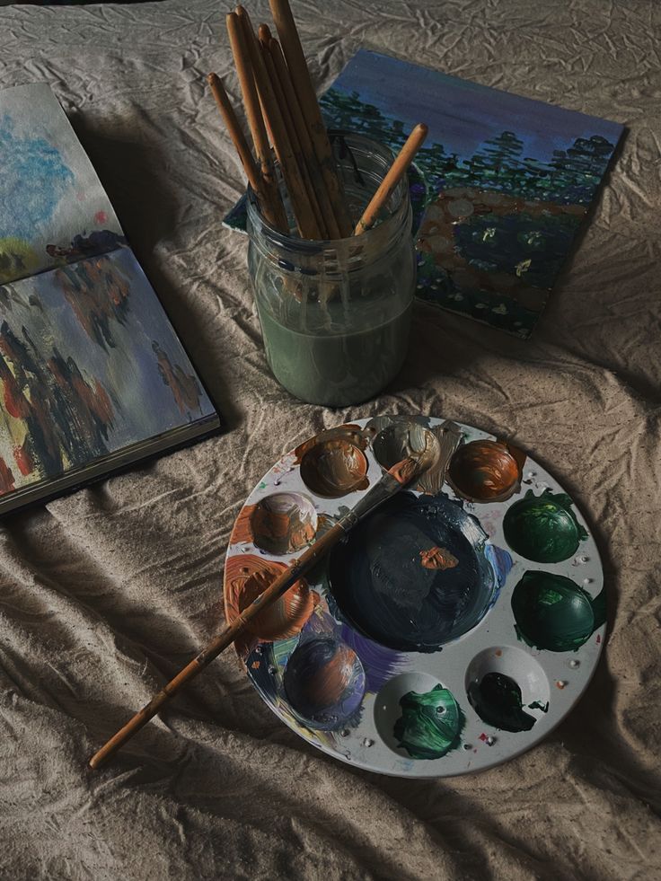 an artist's palette with paints and brushes on a bed next to two paintings