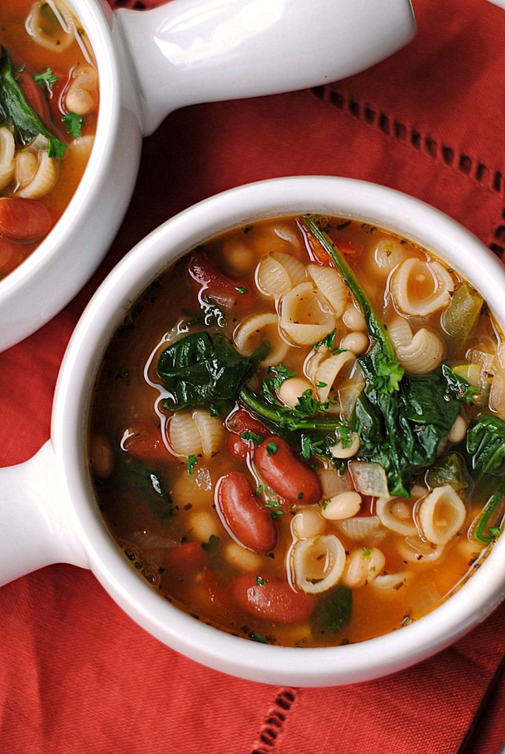 two white bowls filled with pasta and spinach soup on top of a red cloth