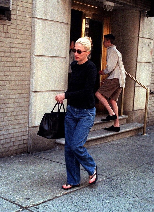 a woman walking down the sidewalk carrying a black bag and wearing sunglasses, with another man in the background