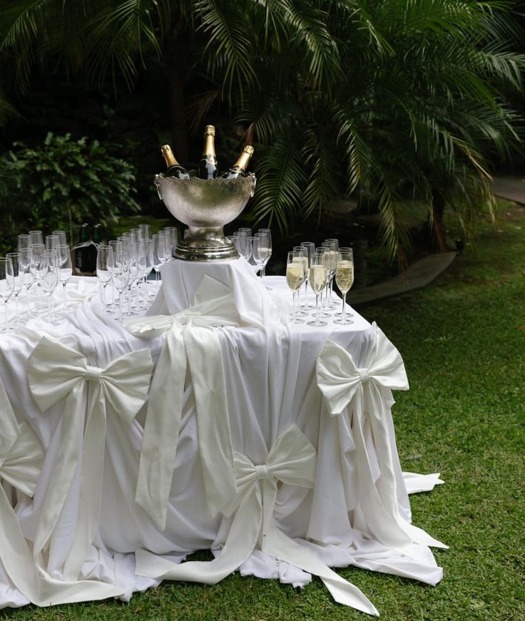 a table covered in white cloths and champagne glasses with gold handles on it, surrounded by palm trees