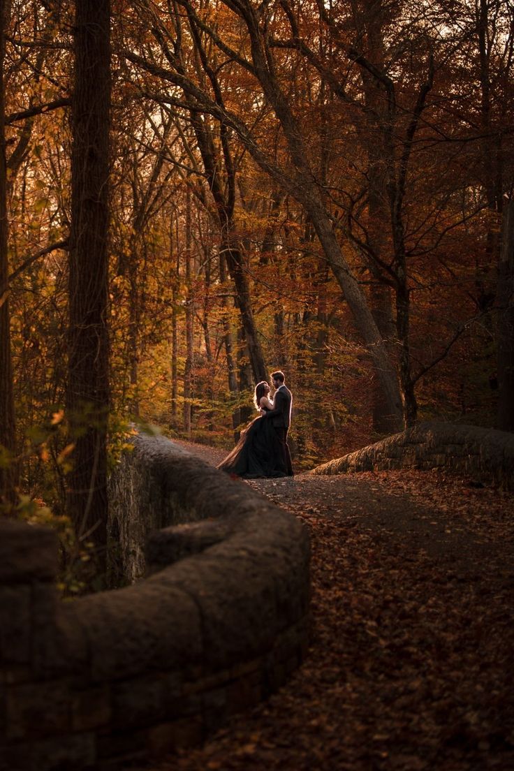 a man and woman are sitting on a stone wall in the middle of an autumn forest