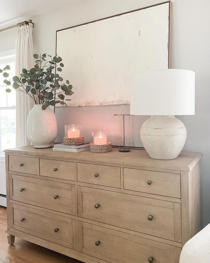 a dresser with two vases on top and candles in front of it, sitting next to a window