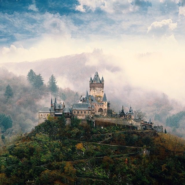 a castle on top of a hill surrounded by trees and fog in the sky with mountains behind it