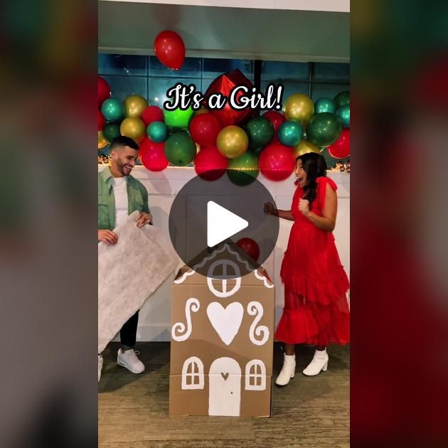 a woman in a red dress standing next to a cardboard gingerbread house with the words it's a girl