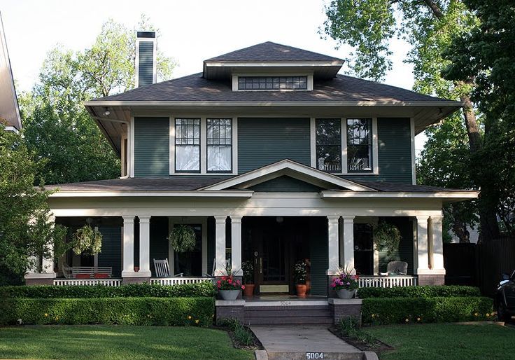 a green house with white trim and pillars on the front porch is surrounded by greenery