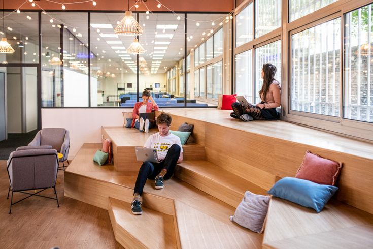 people are sitting on the stairs in an open area with large windows and wooden flooring