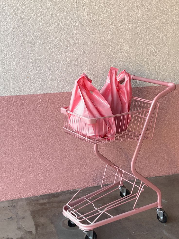 a pink shopping cart filled with bags on top of a cement floor next to a wall