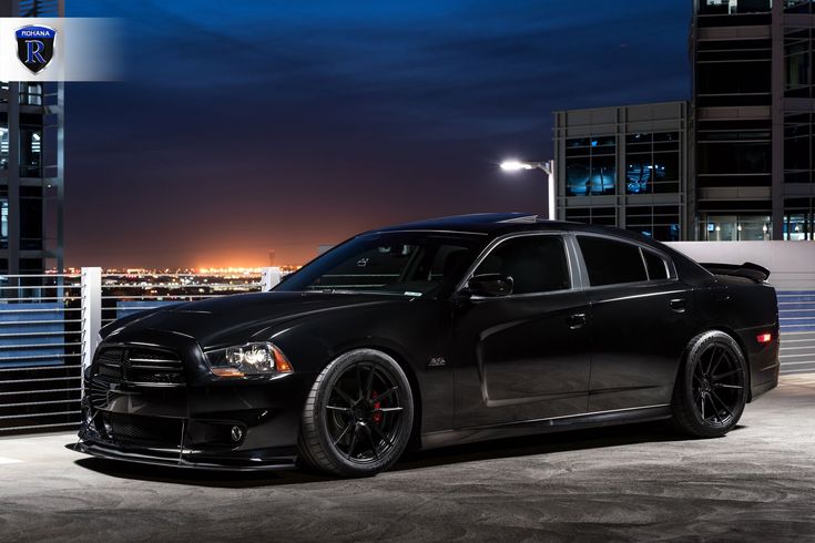 a black dodge charger parked in front of a tall building at night with city lights behind it