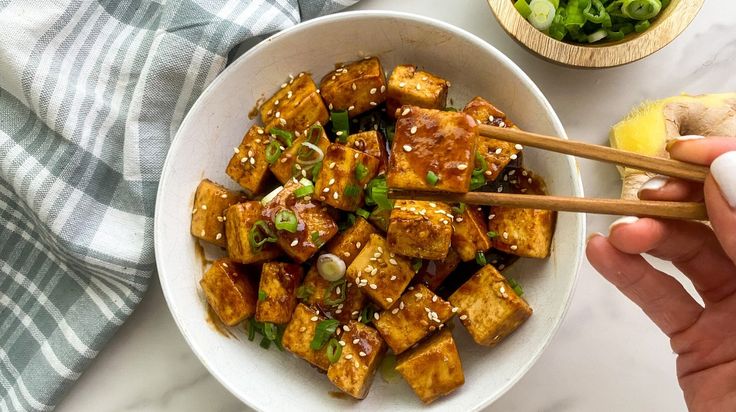 someone holding chopsticks over a bowl of tofu with sauce and green onions